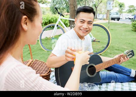 Young happy couple toasting with avec des verres de jus d'orange pendant qu'ils assis sur l'herbe et jouer de la guitare dans le parc Banque D'Images