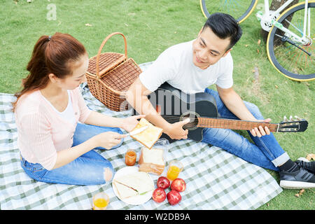 Smiling Young man playing guitar avec sa petite amie, la préparation des toasts et des fruits sur le déjeuner durant leur date à pique-nique dans le parc Banque D'Images