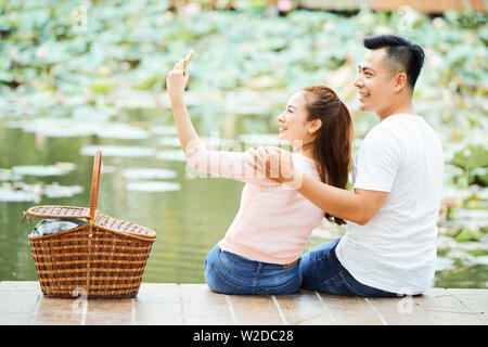 Smiling Asian woman holding mobile phone et décisions portrait selfies avec son petit ami embrasser alors qu'ils assis sur la jetée dans le parc Banque D'Images