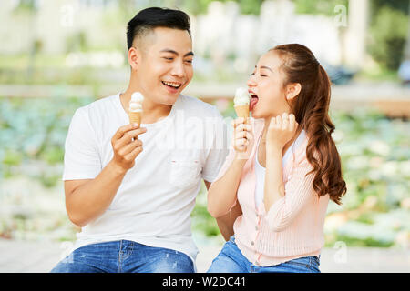 Deux amis asiatiques en vêtements décontractés assis sur un banc et de manger de la crème glacée à l'extérieur doux Banque D'Images