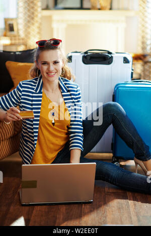Happy voyageur élégant femme en blouson rayé avec carte de crédit l'achat des billets d'avion en ligne sur un ordinateur portable tout en étant assis près de la table et l'sacs trolley Banque D'Images