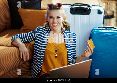 Voyageur seul mode Smiling woman in blouson rayé avec carte de crédit en ligne de réservation des billets d'avion sur un ordinateur portable tout en étant assis près de la table et l'trolle Banque D'Images