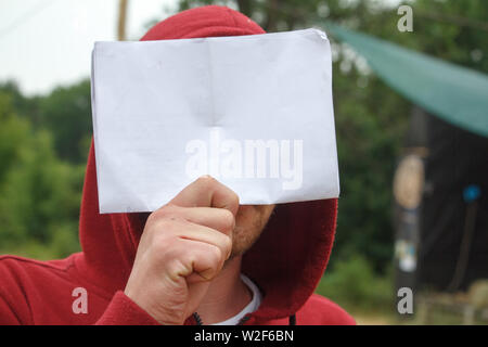 Un jeune homme en rouge hoodie est titulaire d'une feuille de papier blanc en face de son visage, copyspace Banque D'Images