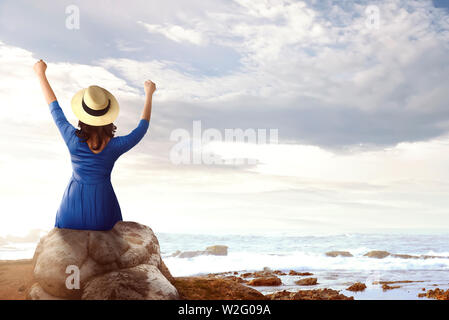 Vue arrière de l'Asian Woman in hat assis sur le rocher et regardant la vue sur l'océan dans la plage Banque D'Images