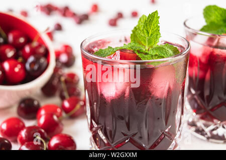 Le jus de cerise douce dans les verres avec les feuilles de menthe et des cubes de glace, et de cerises fraîches sur une table Close Up on White Background Banque D'Images