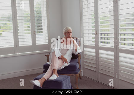 Active senior woman with hand on chin assis sur une chaise dans une maison confortable Banque D'Images