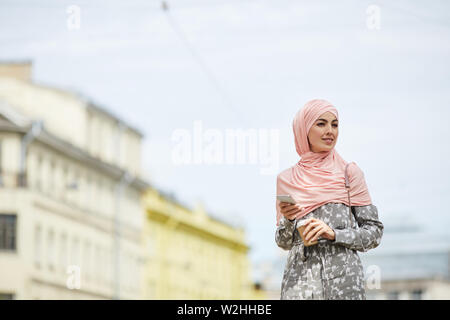 Jeune femme musulmane moderne positive en hijab marcher sur la rue de la ville et de boire du café pendant l'utilisation de smartphone Blackberry Banque D'Images