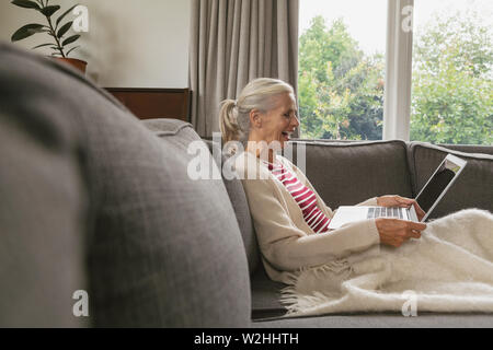 Active senior woman relaxing on sofa and using laptop dans la salle de séjour confortable à l'accueil Banque D'Images