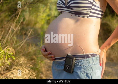 Une femme enceinte diabétique avec une pompe à insuline dans une forêt. Elle porte une jupe bleue et un haut de bikini à rayures. Banque D'Images