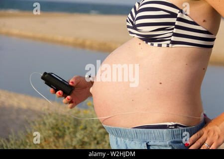 Une femme enceinte diabétique avec une pompe à insuline sur la plage. Elle porte une jupe bleue et un haut de bikini à rayures. Banque D'Images