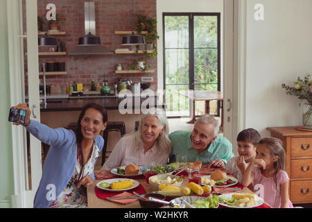 Multi-generation family en tenant avec selfies téléphone mobile tout en ayant de la nourriture sur table à manger Banque D'Images