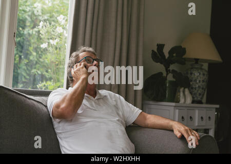 Senior man sitting on sofa and talking on mobile phone dans la salle de séjour confortable à l'accueil Banque D'Images