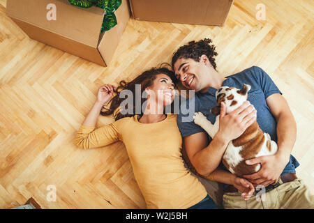 Le jeune couple est heureux de déménager dans une nouvelle maison. Ils sont couchés sur le sol avec leur petit chiot après ils ont apporté des boîtes avec des choses à th Banque D'Images