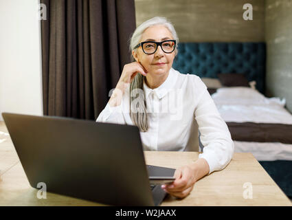 Hauts femme aux cheveux longs portant des lunettes de pointage à l'ordinateur à la table dans son appartement confortable Banque D'Images