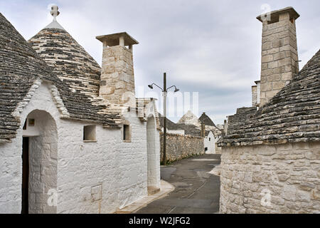 La rue médiévale avec des maisons trulli sur le fond de ciel nuageux dans la ville d'Alberobello en Italie. Il y a de grandes cheminées sur les toits. L'horizontale. Banque D'Images