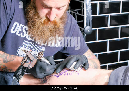 Un tatoueur ou tatoueur travaille sur un client Créer un tatouage ou un morceau de body art en utilisant une machine à tatouer électrique. Banque D'Images