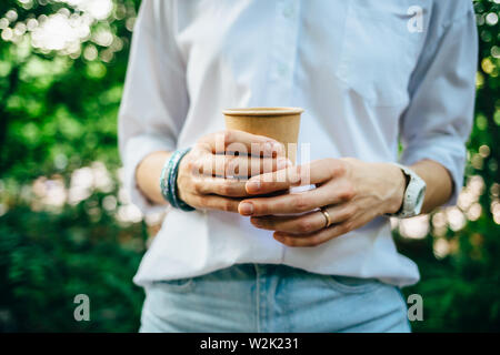 Close-up of female's hands holding brown paper cup. Jeune femme portant une chemise blanche et un jean bleu de boire du café à l'extérieur dans le parc avec green Banque D'Images