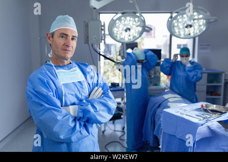 Homme surgeon standing avec bras croisés en salle d'opération alors que les chirurgiens se préparer pour l'opération Banque D'Images