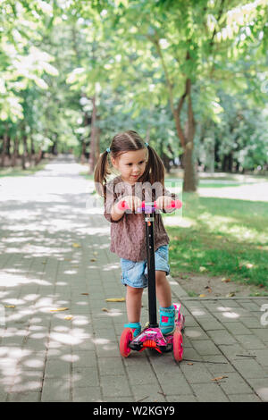 Petit enfant fille apprendre à conduire un scooter dans un parc de la ville aux beaux jours d'été. Banque D'Images