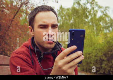 Curieusement, la pensée de l'homme tenant un smartphone, assis dehors dans le jardin. Un jeune gars regarde indignée à l'écran de son smartphone. Mauvaise lecture Banque D'Images