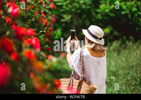 Femme de tourisme en chapeau de paille et des vêtements blancs photoshoots rose fleur sur smartphone. Banque D'Images