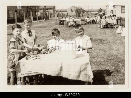 La photographie des enfants de l'école primaire des années 1960 portant des tabliers ou des bavoirs, jouant avec de la pâte à jouer sur des tables disposées dans les terrains de jeu de l'école, à l'extérieur, à l'extérieur, peut-être Leicestershire, U.K. Banque D'Images