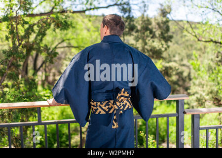 Jeune homme en costume kimono debout dos leaning on railing clôture dans un jardin extérieur avec vue sur la nature au Japon Banque D'Images