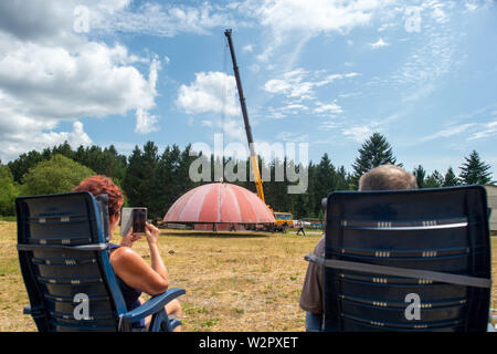 Benneckenstein, Allemagne. 10 juillet, 2019. Une grue soulève le toit d'un ancien dôme radar sur le terrain de l'Ostdeutsche Fahrzeug- und Technikmuseum afin de le soulever sur les murs de l'établissement. Au premier plan, passionnés de technologie s'asseoir dans des chaises longues. Le dôme radar se tenait sur le Brocken jusqu'au début des années 90 et à l'abri des installations techniques militaires sensibles. Le 13 juillet 2019 et 14 juillet 2019 le 4e jours de la technologie auront lieu au Musée de la technologie. Puis le dôme sera visible. Credit : Klaus-Dietmar Gabbert/dpa-Zentralbild/ZB/dpa/Alamy Live News Banque D'Images