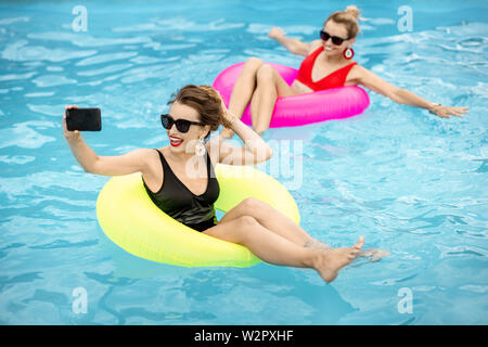 Faire copines avec photo selfies téléphone tout en nageant à anneaux gonflables dans l'eau chauffée à l'extérieur pendant l'été Banque D'Images