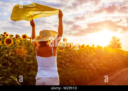 Happy senior woman walking in blooming champ de tournesol élever les mains à l'écharpe et s'amusant. Vacances d'été. Banque D'Images