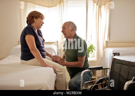 Femelle adulte patient assis sur le lit d'hôpital, son mari à genoux à côté d'elle tenant sa main. Banque D'Images