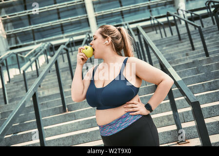 Alimentation saine. Grande taille femme en vêtements de sport manger vert apple tout en se tenant debout sur l'escalier extérieur. Le bien-être. Concept de santé. La perte de poids Banque D'Images