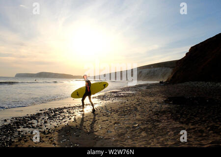 Un jeune homme portant sa planche de surf sous le bras tête dans la mer à pied avec le soleil couchant et Tennyson en bas des falaises de craie dans l'arrière-plan