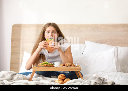 Jeune femme d'une dépendance à l'alimentation manger de savoureux burger on bed at home Banque D'Images