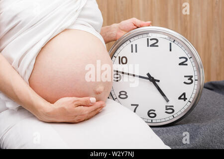 Pregnant woman holding her belly au cours des dernières semaines de la grossesse assis par réveil Banque D'Images