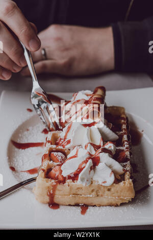 Close up of male hands holding une fourchette, manger une gaufre avec de la crème fraîche et sauce aux fraises, selective focus. Banque D'Images