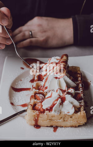 Close up of male hands holding une fourchette, manger une gaufre avec de la crème fraîche et sauce aux fraises, selective focus. Banque D'Images
