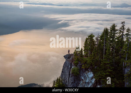 Female Hiker au sommet d'une montagne couverte de nuages pendant un été animé coucher du soleil. Pris sur le dessus de la Sommet, West Vancouver (Colombie-Britannique) Banque D'Images