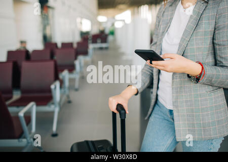 Jeune fille à l'aéroport, debout avec les bagages, l'attente pour le départ. Holding passport et billet. Téléphone cellulaire de défilement Banque D'Images