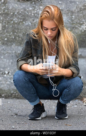 Les jeunes en jeans femme accroupi elle écoute de la musique sur son téléphone mobile perdu dans la sélection d'un morceau à jouer Banque D'Images