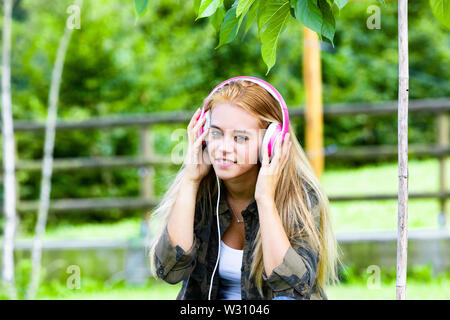 Smiling attractive young woman enjoying sa musique sur un casque stéréo alors qu'elle se détend à l'extérieur dans un jardin ou le parc. Banque D'Images