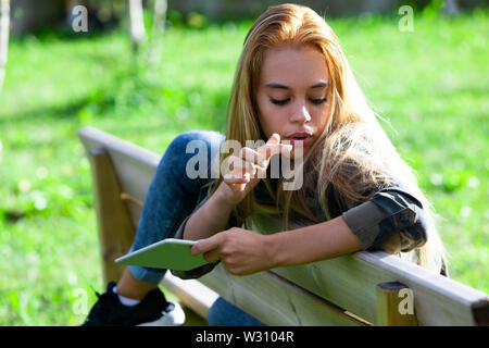 Jeune femme de détente en plein air sur un banc de parc avec sa jambe drapé sur le haut de la holding a tablet regardant quelque chose sur ses longs cheveux avec le doigt sur le point Banque D'Images