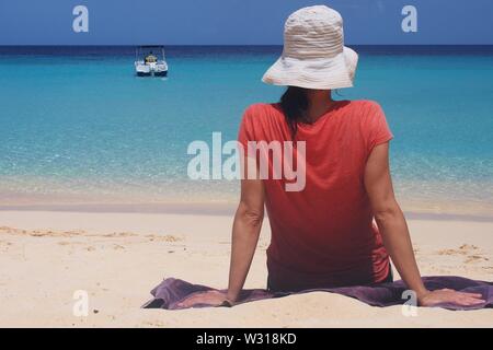 Dos d'une femme assise sur une plage, face à la mer. White Hat et Red t-shirt. Meads Bay, Anguilla, BWI. Un hors-bord visible dans la distance. Banque D'Images