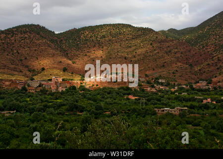 Vue sur la montagne et les maisons des villages berbères dans les montagnes de l'Atlas du Maroc en Afrique du Nord, montrant robuste vert arbres, buissons et paysage de boue rouge Banque D'Images