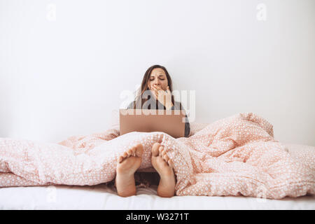 Les jeunes adultes belle fille bénéficie d'un ordinateur portable dans le lit et bâille. Elle était fatiguée ou s'est réveillé tôt et somnolent. Banque D'Images