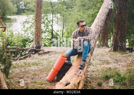 Jeune homme s'asseoir sur le camion en forêt et à l'aide de téléphone mobile. l'homme est assis sur un grand voyageur arbre tombé, holding phone dans la main thème voyage nature randonnée. Banque D'Images