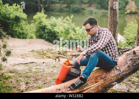 Jeune homme s'asseoir sur le camion en forêt et à l'aide de téléphone mobile. l'homme est assis sur un grand voyageur arbre tombé, holding phone dans la main thème voyage nature randonnée. Banque D'Images