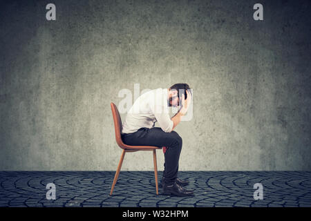 L'homme déprimé contrarié par la mauvaise chance assis sur une chaise, regardant vers le bas contre mur gris Banque D'Images