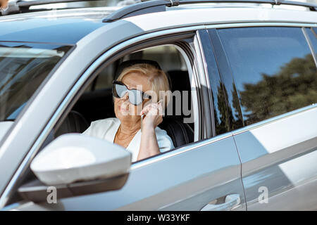 Belle senior business woman avec téléphone en conduisant une voiture moderne. Concept d'une population active au cours de l'âge de la retraite Banque D'Images