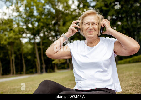 Happy senior femme à écouter la musique avec un casque tout en pratiquant le yoga dans le parc. Concept d'une vie active à la retraite Banque D'Images